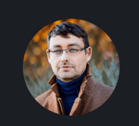 Man with glasses and a coat, standing outdoors with autumn leaves in the background.
